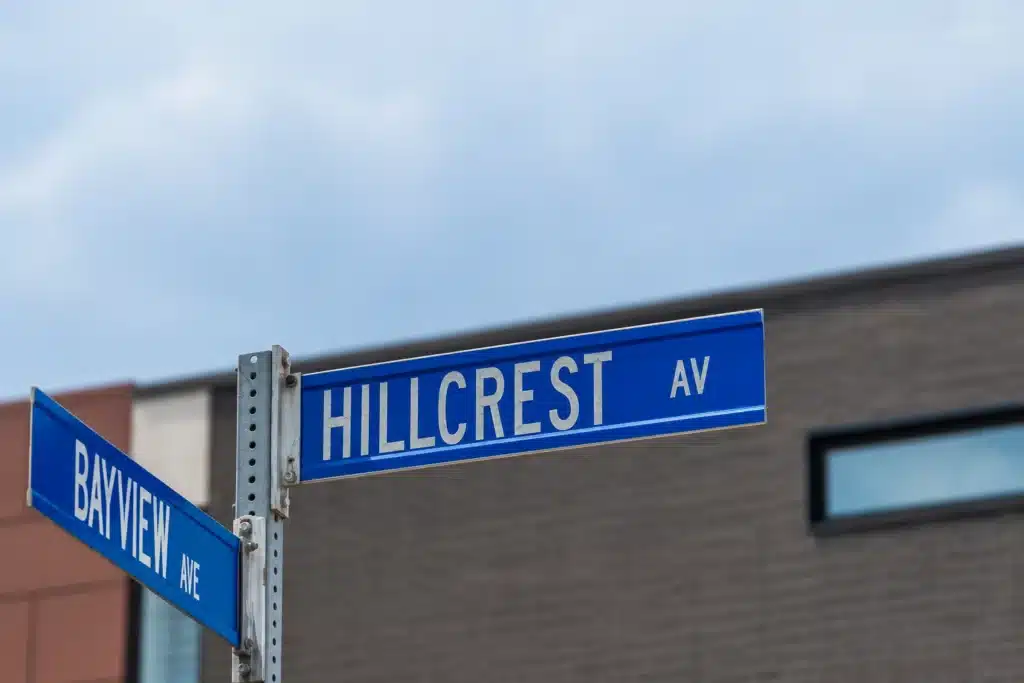 Blue Hillcrest Avenue and Bayview Avenue street signs at an intersection, with a modern building and cloudy sky in the background.