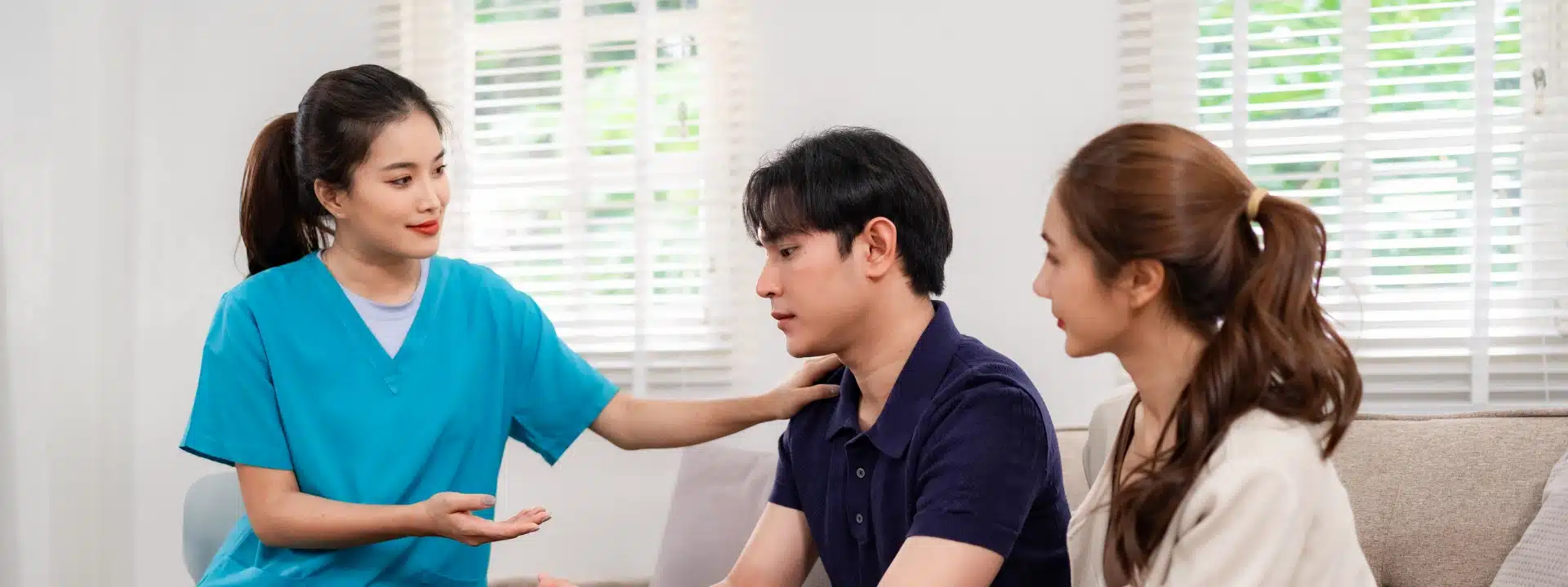 Asian woman converses with a man and woman seated on a couch in a cozy living room setting.